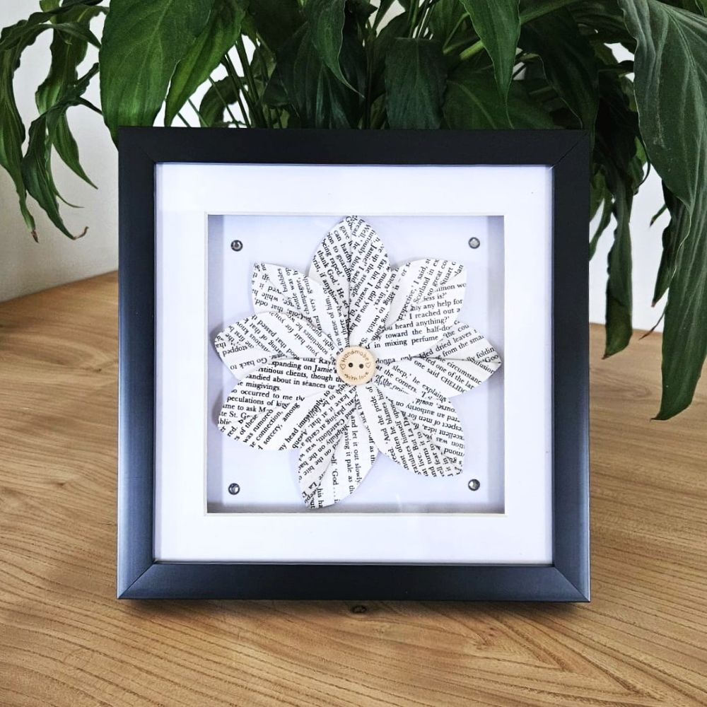 Framed paper flower art on a wooden surface with a plant in the background