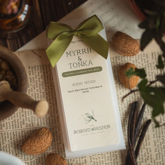 A boxed wax melt set labelled 'MYRRH & TONKA' by Robins Whisper, tied with an olive green ribbon, photographed in a styled flat-lay with spices, vanilla beans, and printed paper.