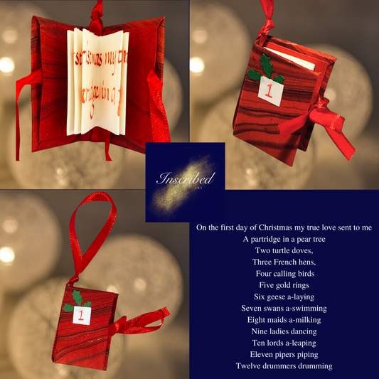 A set of three miniature book Christmas decorations with red watercolor illustrations and text, displayed against a festive background with Christmas lights.
