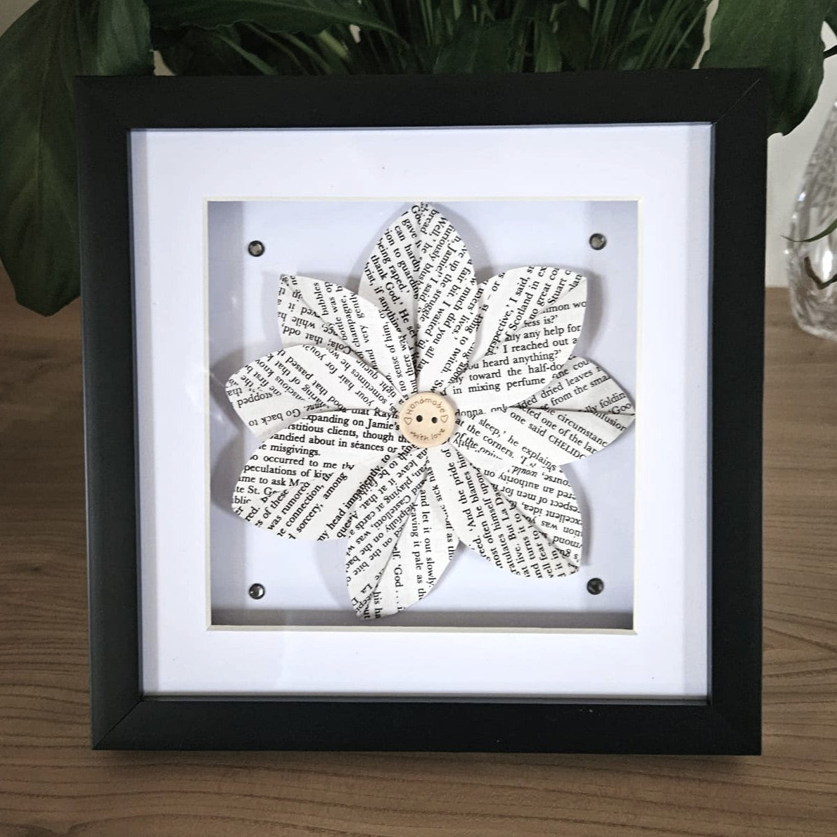 Decorative flower made from book pages in a black frame on a wooden surface with plants in the background.