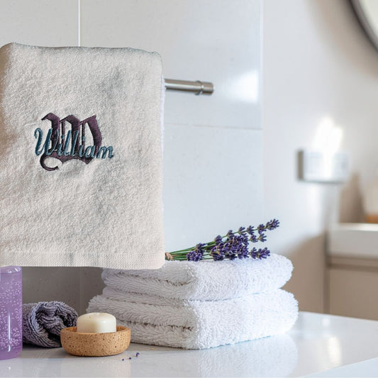 White towel with embroidered name, stacked towels, and lavender on a bathroom counter.