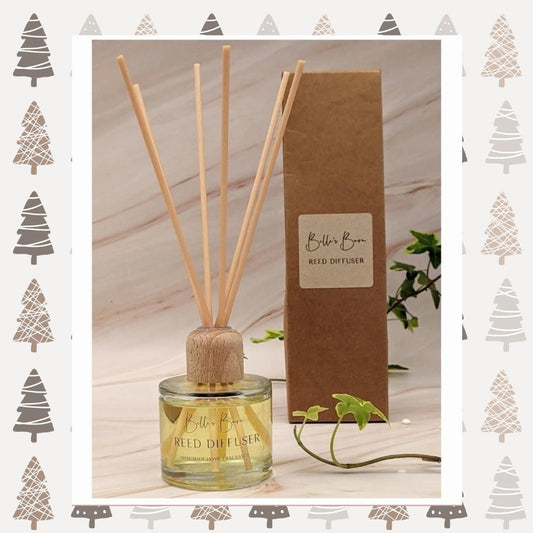 A reed diffuser with a wooden lid on top of a round glass bottle, accompanied by six reeds. The diffuser is labeled with 'Belle's Barn' and 'Reed Optimiser' on a kraft gift box background with Christmas-themed decorations in the corners.