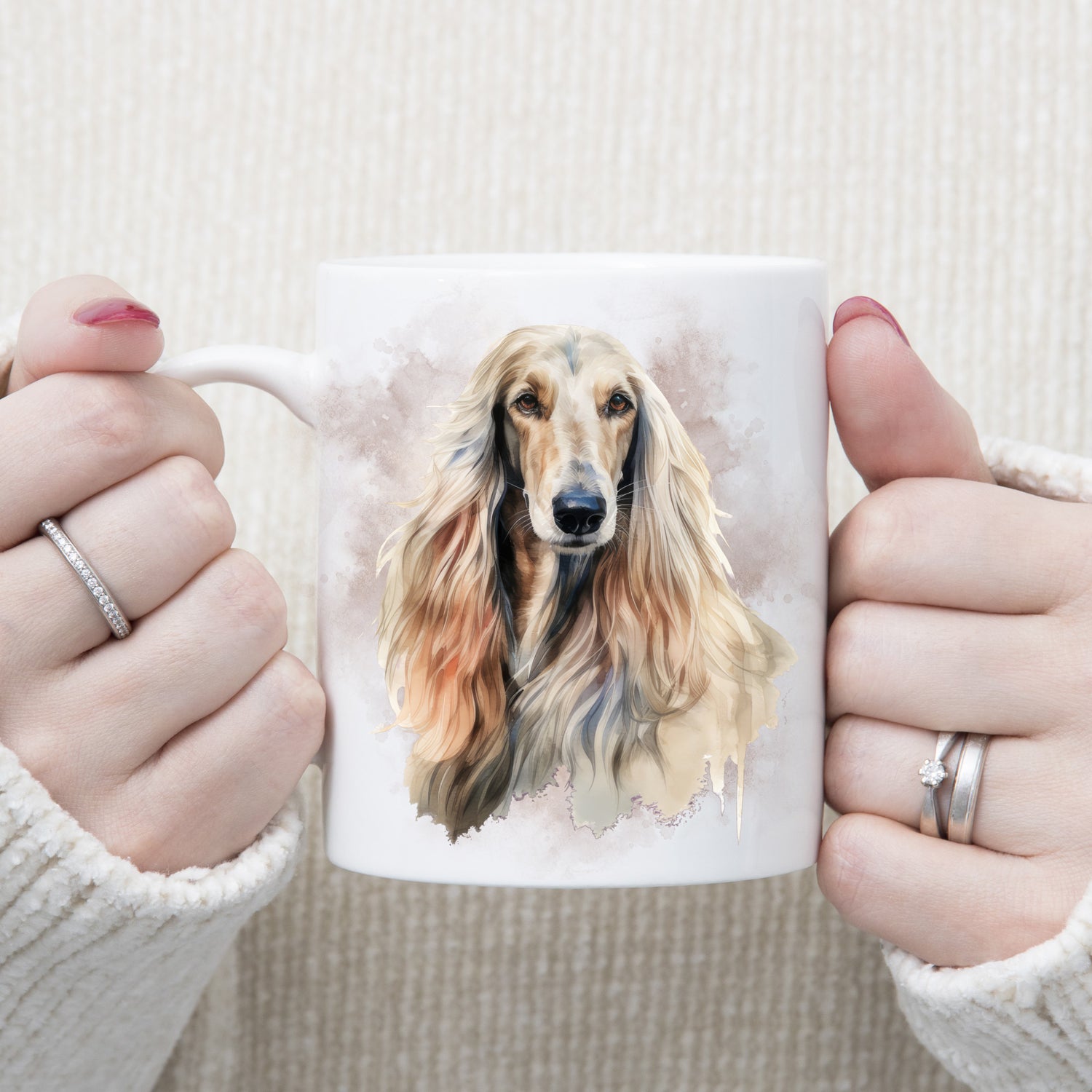 A white ceramic mug with a head image of an Afghan Hound with long flowing hair.  A grey/brown smoke effect decorates the background.  A woman is holding the mug in two hands.
