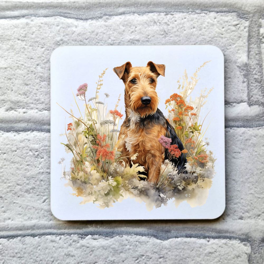 An Airedale Terrier is sat among multi-coloured  wildflowers looking towards the right.  This image is on a  white coaster.    The coaster is placed on a grey, brick effect surface.