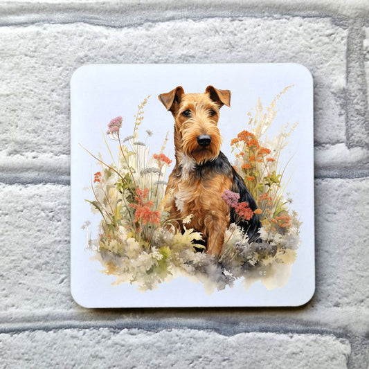 An Airedale Terrier is sat among multi-coloured wildflowers looking towards the right. This image is on a white coaster. The coaster is placed on a grey, brick effect surface.