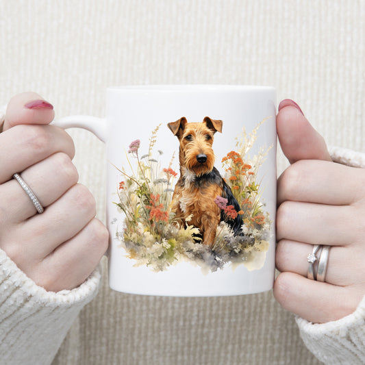 An Airedale Terrier dog is in a bed of orange and purple wildflowers on a white ceramic mug. The mug is being held by a woman with both hands.
