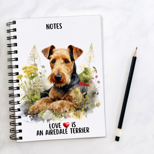 White spiral bound notebook with an Airedale Terrier laid among wildflowers on the front and a quote written at the bottom. A black pencil is placed to the right hand side.