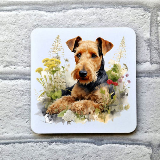 An Airedale Terrier is laid in a bed of various plants and flowers. This image is on a white coaster. The coaster is placed on a grey, brick effect surface.