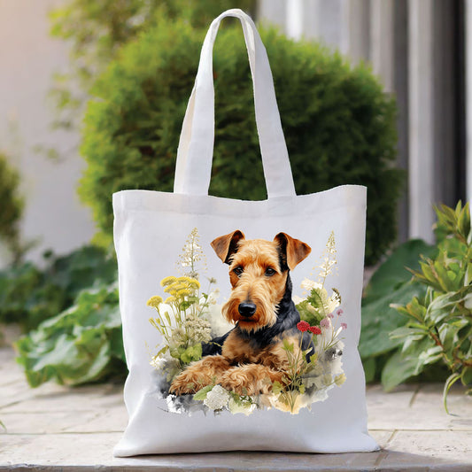 White Airedale Terrier tote bag with a long handle. Red and yellow flowers decorate. The bag is placed in front of a large green shrub with green foliage to the right side.