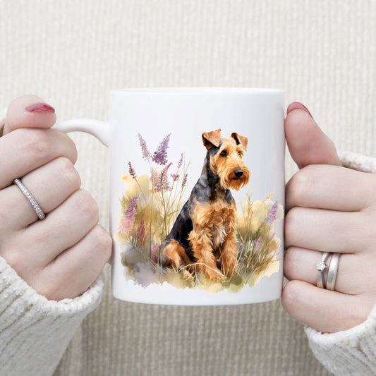 An Airedale Terrier dog is sitting in a grassy field with purple flowers on a white ceramic mug. A woman is holding the mug in two hands.