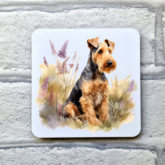 An Airedale Terrier is sat among grass and purple pointed flowers looking towards the right. This image is on a white coaster. The coaster is placed on a grey, brick effect surface.