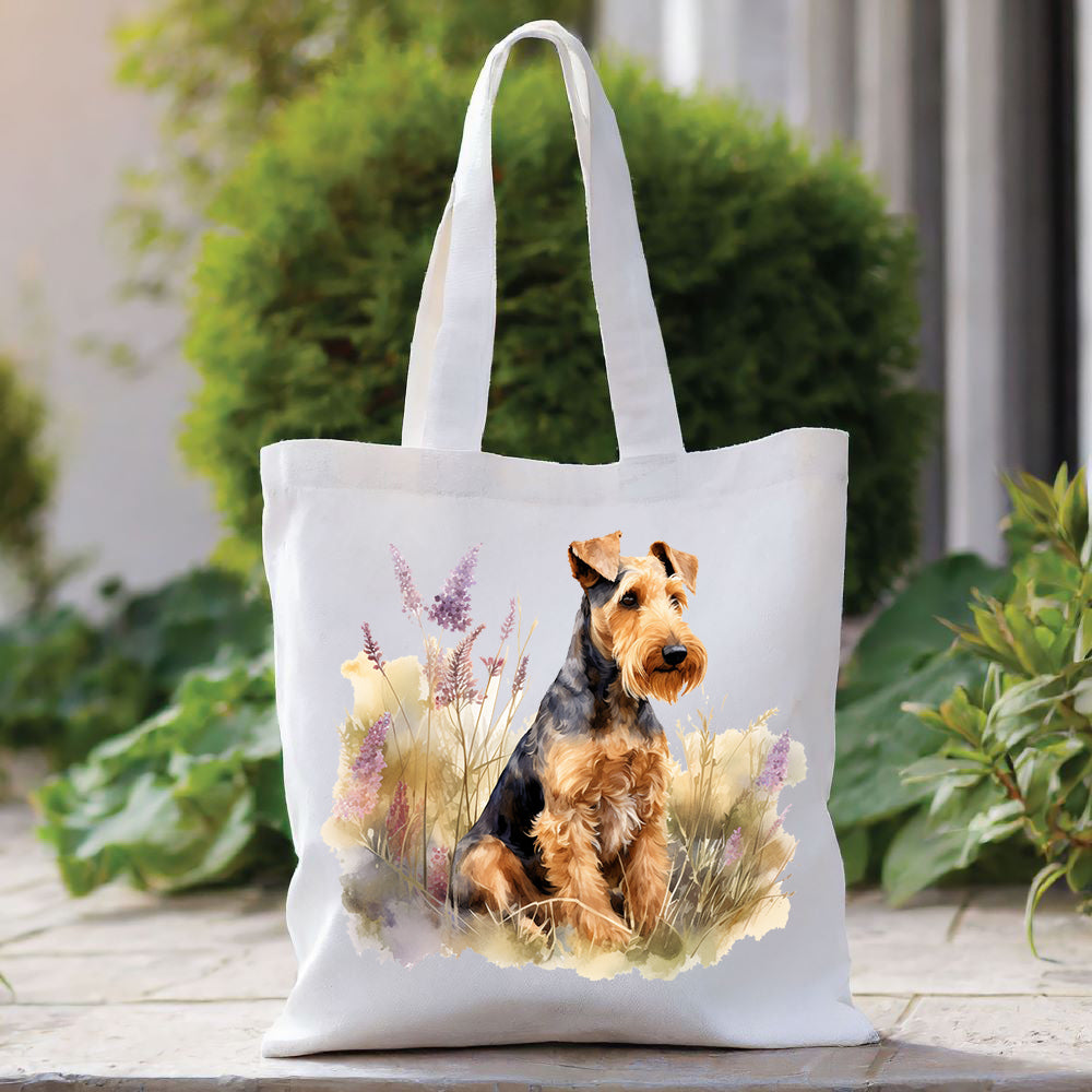 White tote bag with a long handle shows an Airedale Terrier sat among tall purple flowers and looking toward the right.  The bag is placed in front of a large green shrub with green foliage to the right side.