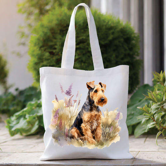 White tote bag with a long handle shows an Airedale Terrier sat among tall purple flowers and looking toward the right. The bag is placed in front of a large green shrub with green foliage to the right side.