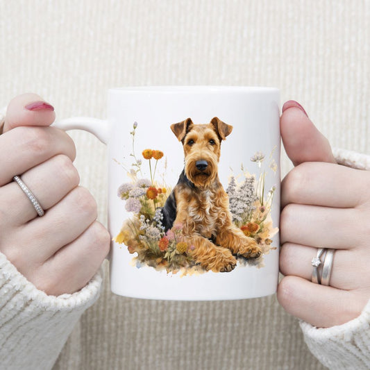 An Airedale Terrier dog is laid among a bed of orange and white wildflowers on a white ceramic mug. A woman is holding the mug with both hands.