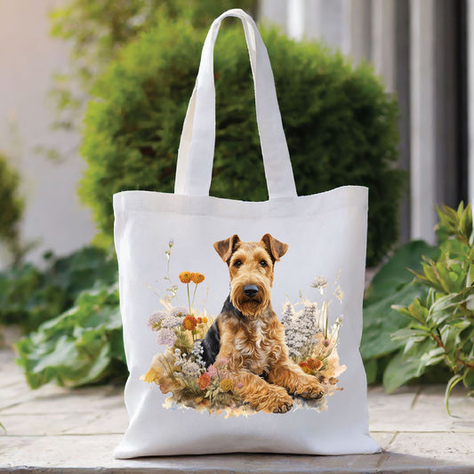 White tote bag with a long handle shows an Airedale Terrier laid among a flower bed and looking forward. The bag is placed in front of a large green shrub with green foliage to the right side.