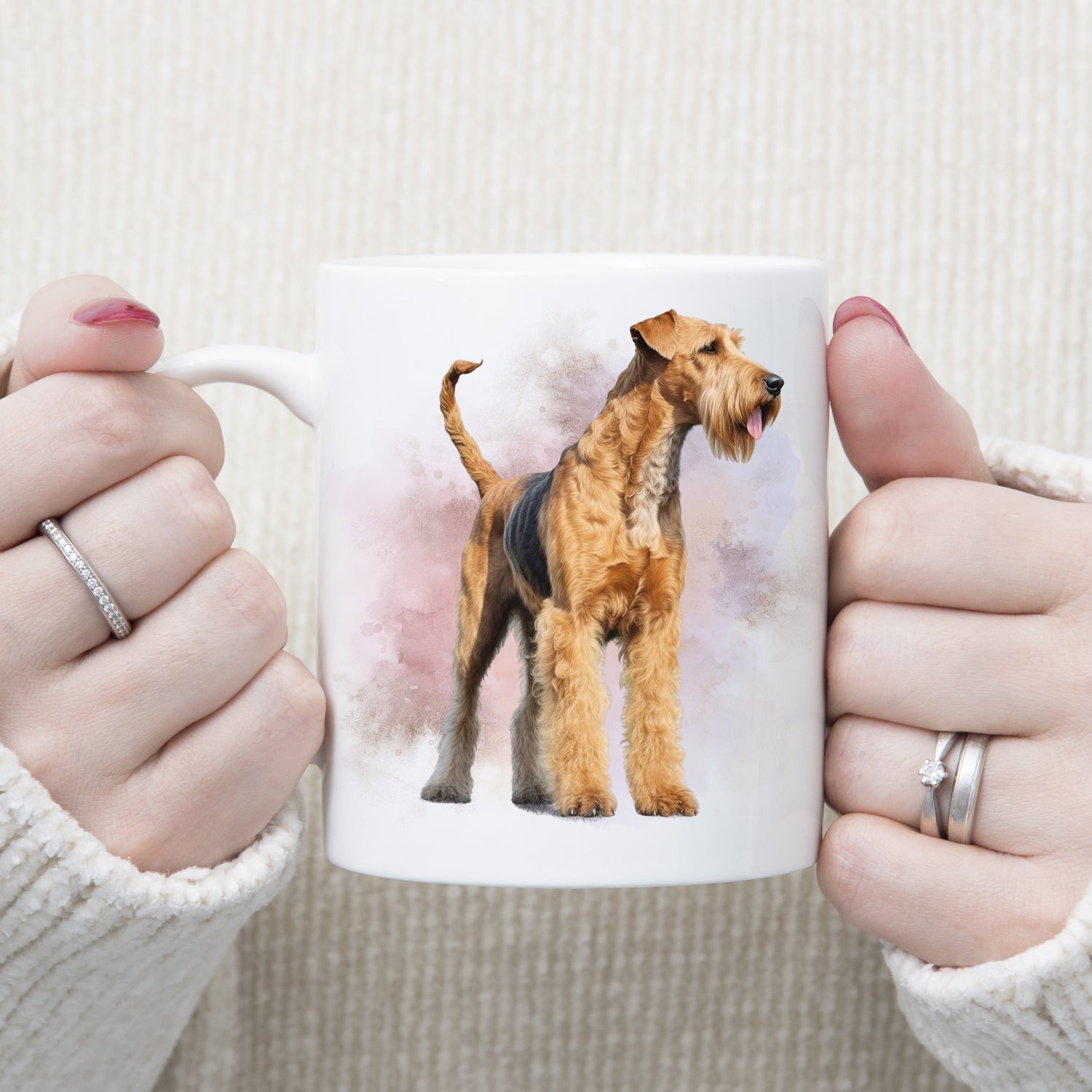 An Airedale Terrier dog is stood facing right in front of a smoky background on a  white ceramic mug. The mug is being held by a woman with both hands.