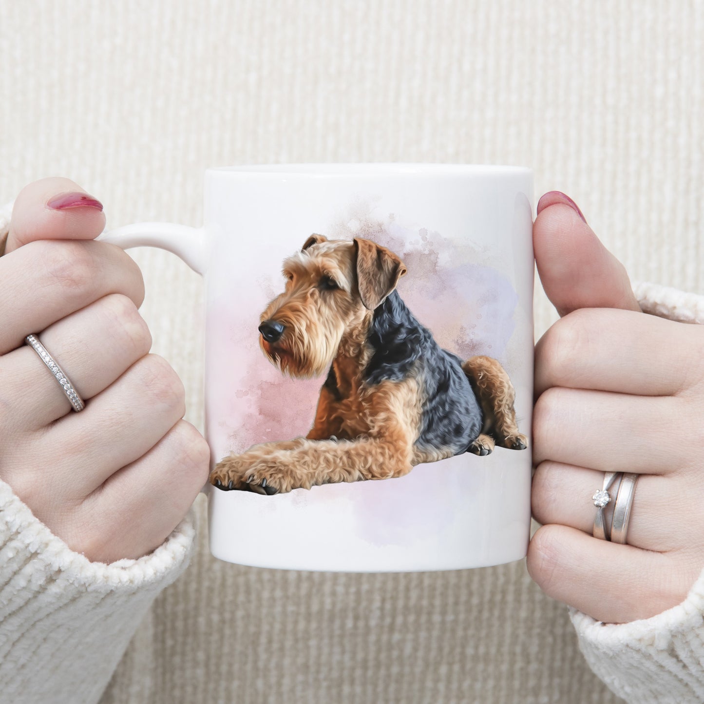 An Airedale Terrier dog is laid with a pink and lilac smoky background on a  white ceramic mug. A woman is holding the mug with both hands.