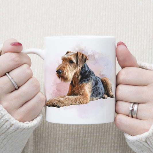 An Airedale Terrier dog is laid with a pink and lilac smoky background on a white ceramic mug. A woman is holding the mug with both hands.