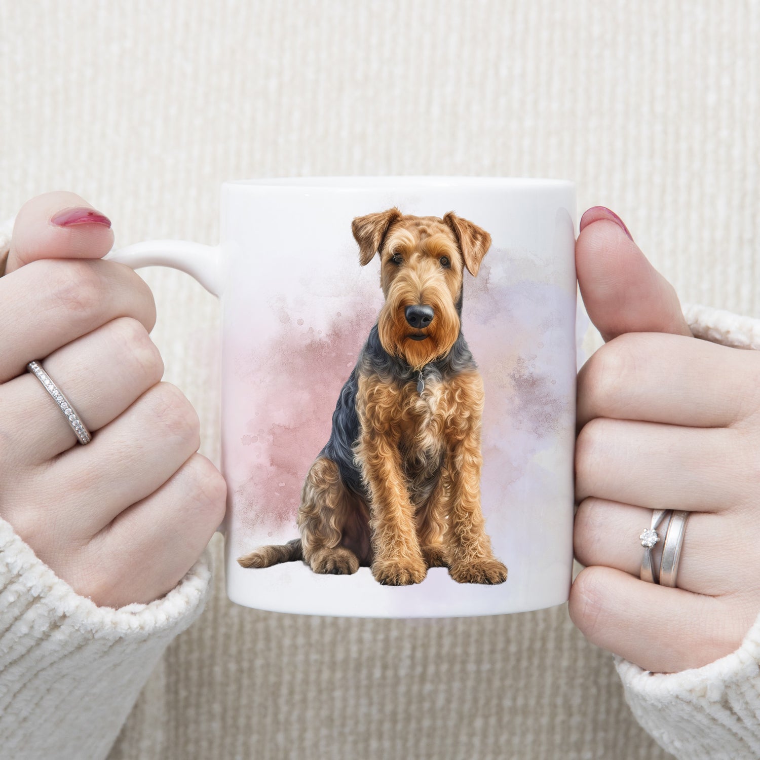 An Airedale Terrier dog is sat in front of a pink and lilac smoky background on a  white ceramic mug. A woman is holding the mug with both hands.