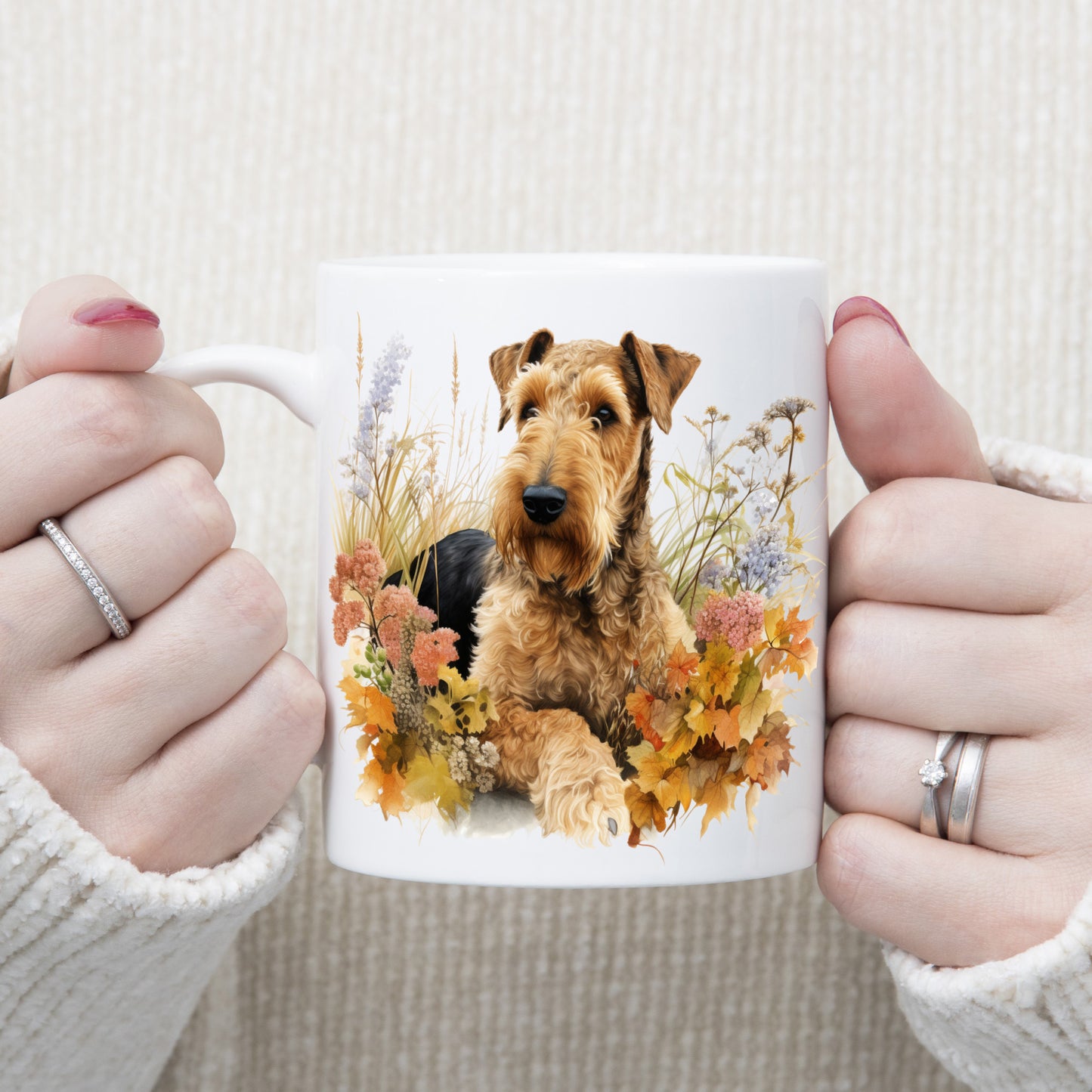 An Airedale Terrier is laid among a bed of Autumnal plants and flowers on a  white ceramic mug. The mug is being held by a woman with both hands.