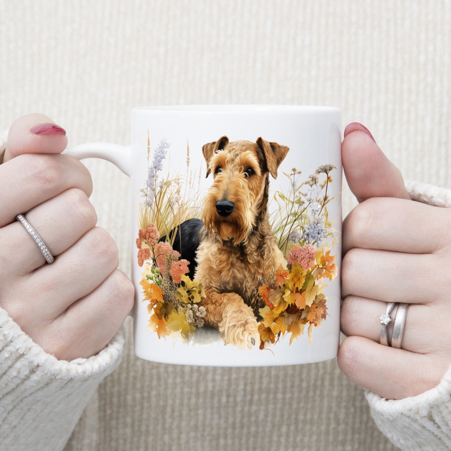 An Airedale Terrier is laid among a bed of Autumnal plants and flowers on a  white ceramic mug. The mug is being held by a woman with both hands.