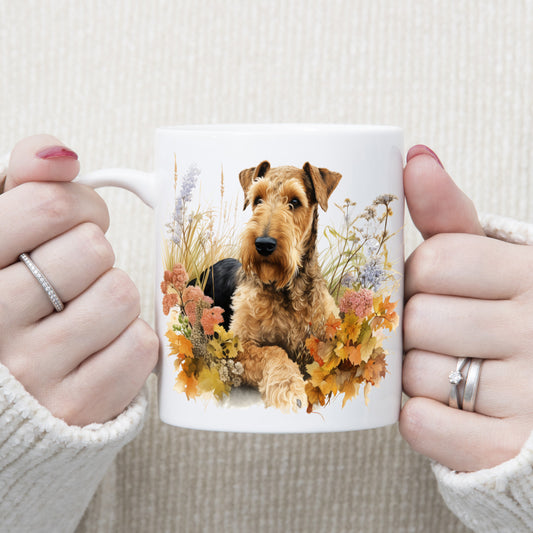 An Airedale Terrier is laid among a bed of Autumnal plants and flowers on a white ceramic mug. The mug is being held by a woman with both hands.
