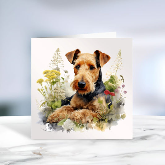 Airedale Terrier laid among various plants and facing forward on a white card.  The card is placed on a grey marble surface.