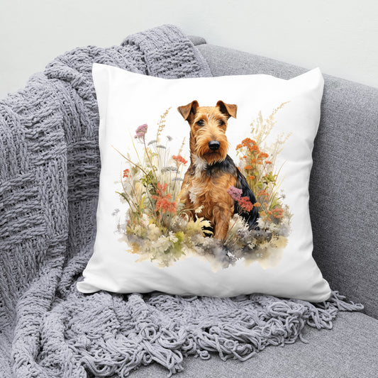 Airedale Terrier sat among wildflowers and facing right on a white cushion.  The cushion is placed in the corner of a grey sofa with a grey blanket draped across the corner.
