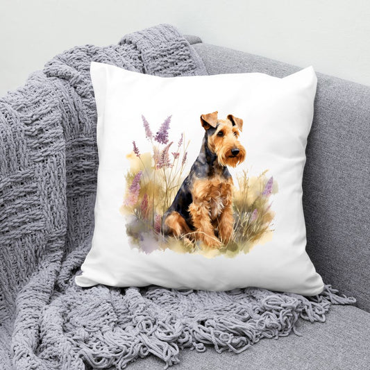 Airedale Terrier sat among purple flowers and facing right on a white cushion.  The cushion is placed in the corner of a grey sofa with a grey blanket draped across the corner.