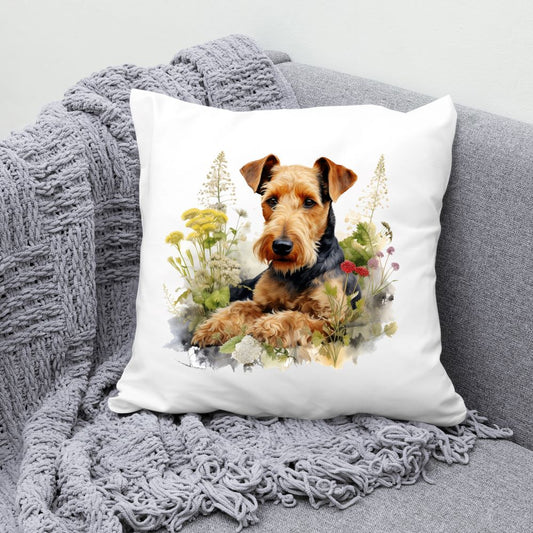 Airedale Terrier laid among various flowers and facing forward on a white cushion.  The cushion is placed in the corner of a grey sofa with a grey blanket draped across the corner.