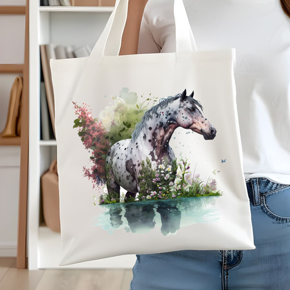 White tote bag with a long handle showing an Appaloosa  horse standing in water with wildflowers.  A woman is holding the bag  in front of white book case.