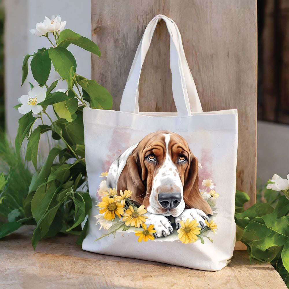 White tote bag with a long handle shows a red and white Basset Hound laid with its head on its paws among yellow flowers and looking forward.  The bag is stood against a wood panel.  Green foliage and white flowers decorate.