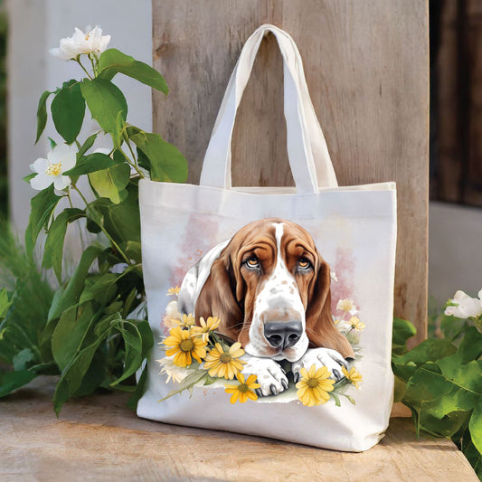 White tote bag with a long handle shows a red and white Basset Hound laid with its head on its paws among yellow flowers and looking forward. The bag is stood against a wood panel. Green foliage and white flowers decorate.