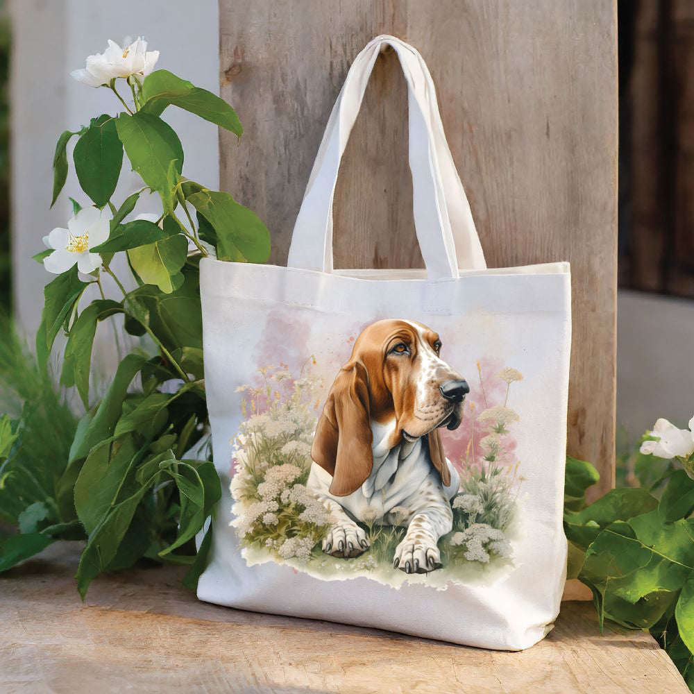 White tote bag with a long handle shows a red and white Basset Hound laid among white flowers and looking toward the right  The bag is stood against a wood panel.  Green foliage and white flowers deco