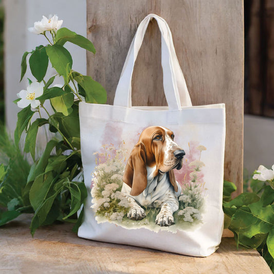 White tote bag with a long handle shows a red and white Basset Hound laid among white flowers and looking toward the right The bag is stood against a wood panel. Green foliage and white flowers deco