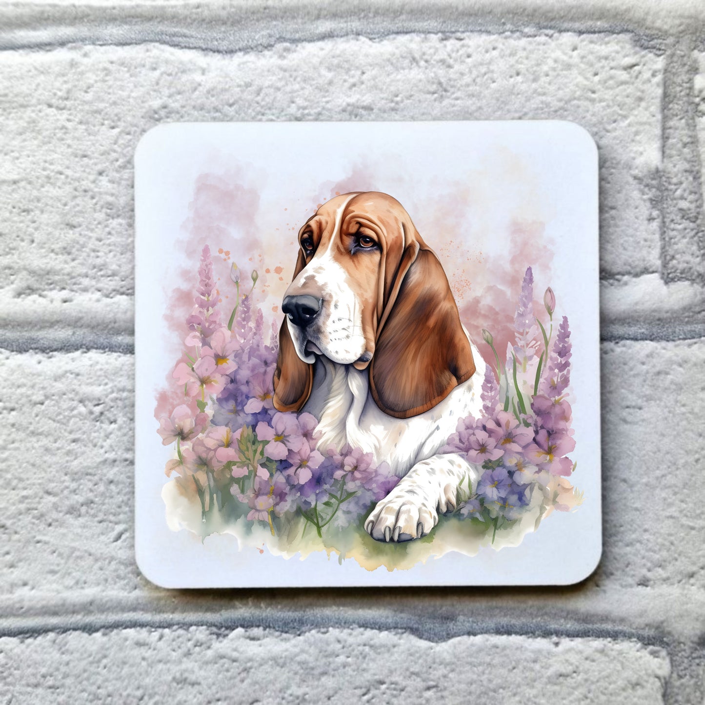 A white table coaster with a  tan and white Basset Hound laid  among purple flowers.  The coaster is placed on a grey, brick effect surface.