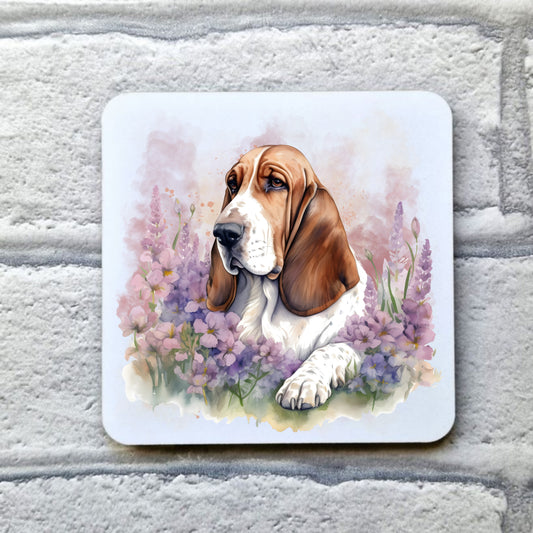 A white table coaster with a tan and white Basset Hound laid among purple flowers. The coaster is placed on a grey, brick effect surface.