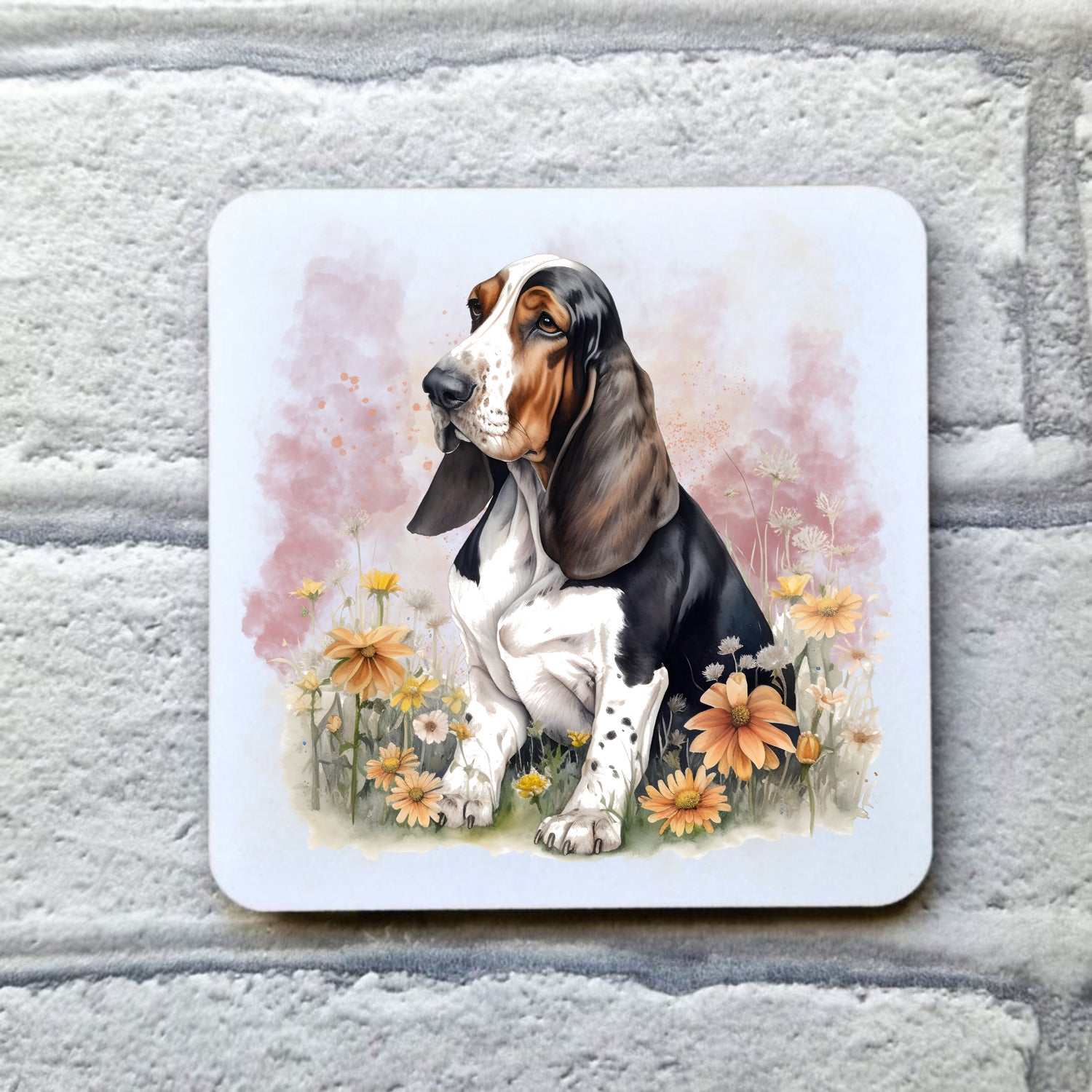 White drinks coaster with a  Tri-coloured Basset Hound sat among orange flowers with a smoky pink background.  The coaster is placed on a grey, brick effect surface.