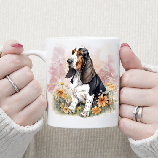 A tri-colour Basset Hound sits facing left among orange flowers.  A pink smoky background decorates this white ceramic mug.  The mug which is being held by a woman with both hands.