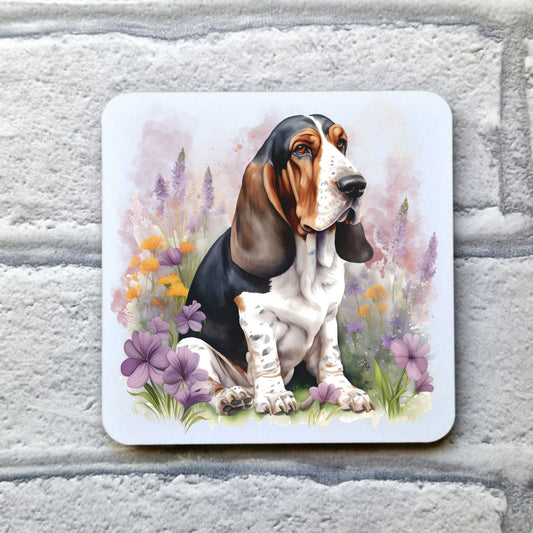A Tri-coloured Basset Hound sat among purple and orange flowers on a white table coaster. The coaster is placed on a grey, brick effect surface.
