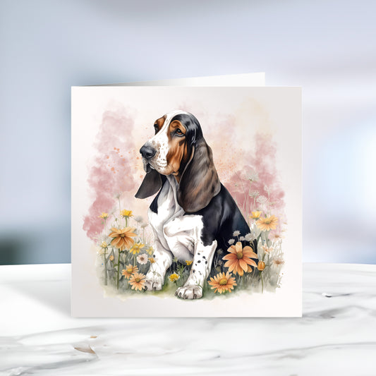 Basset Hound sat among flowers and facing left on a white card or notelet with a smokey pink background.  Card is sitting on a marble surface.