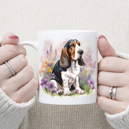 A Basset Hound is sat among orange and purple flowers on a white ceramic mug. A pink smoke effect decorates the background. A woman wearing rings is holding the mug in two hands.