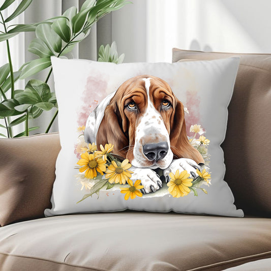 Basset Hound laid among yellow flowers and facing forward on a white cushion.  The cushion is placed in the corner of the sofa with a green plant behind.