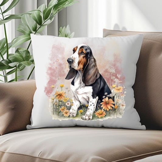 Basset Hound sat among flowers and facing left on a white cushion.  The cushion is placed in the corner of the sofa with a green plant behind.