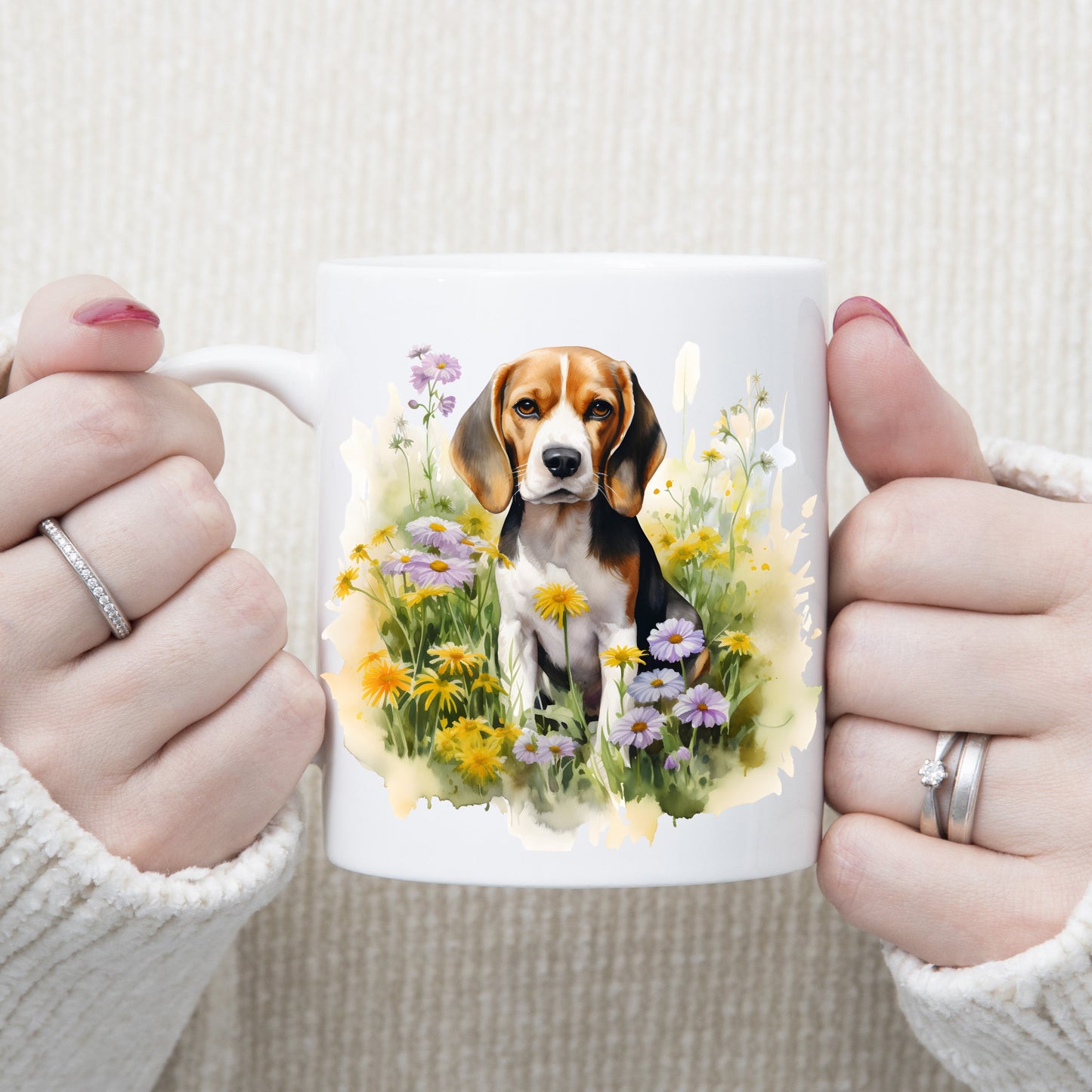 A Beagle dog is sat among purple and yellow wildflowers on a  white ceramic mug. A woman is holding the mug with both hands.
