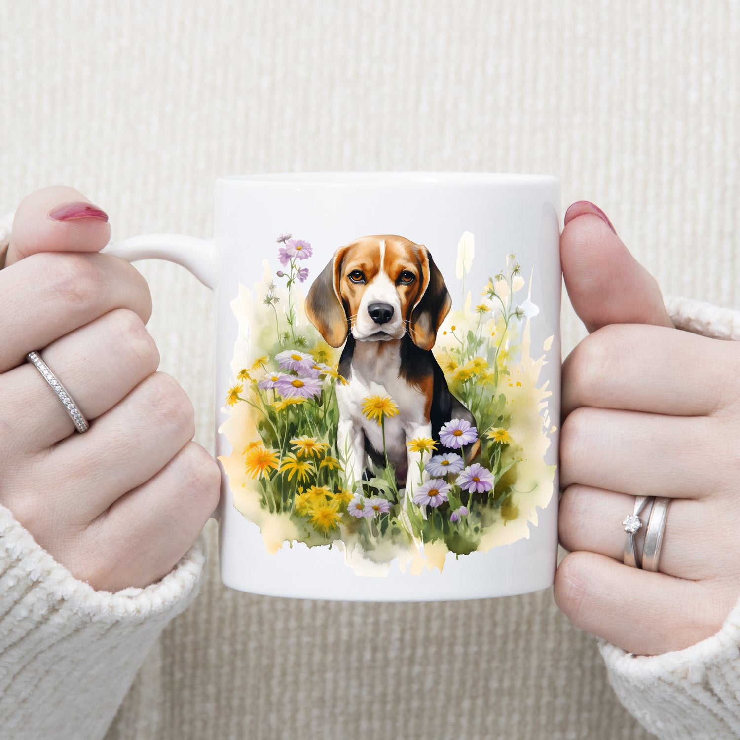 A Beagle dog is sat among purple and yellow wildflowers on a  white ceramic mug. A woman is holding the mug with both hands.