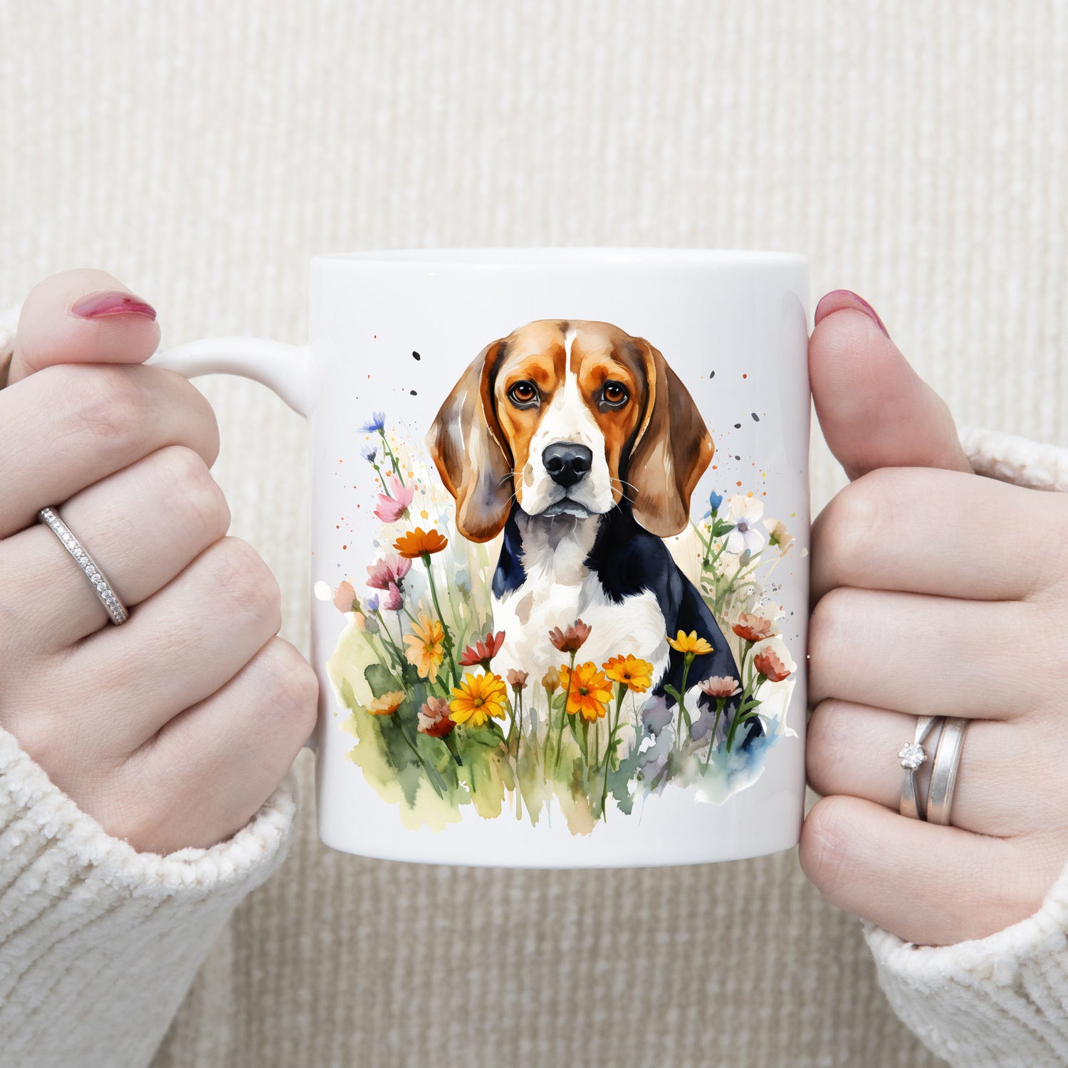 A Beagle dog is sat facing forward among orange daisy-like wildflowers on a  white ceramic mug. A woman is holding the mug with both hands.