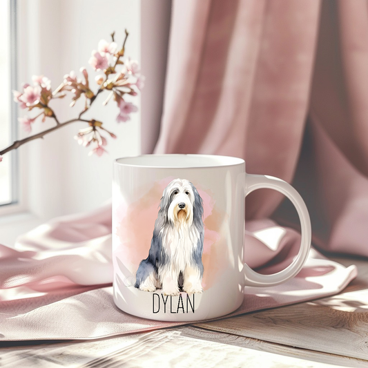 Bearded Collie in a sitting position on a white ceramic mug which has been placed on a pink drape. A stem of small pink flowers accessorizes the image.