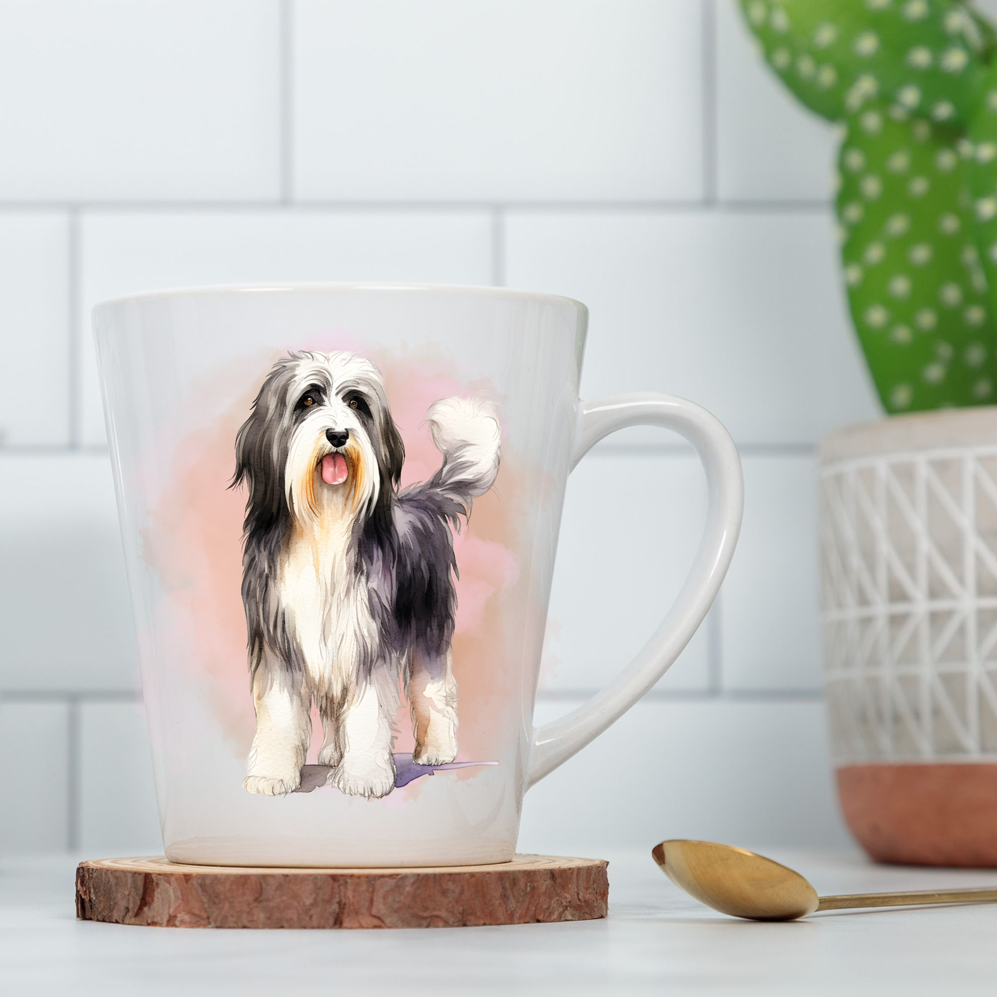 A Bearded Collie is shown standing on a white ceramic latte mug in a pink misty background.  The mug is placed on a wooden disc with a green cactus plant in a decorative pot to the right hand side.  The scene is set against a white tiled wall.