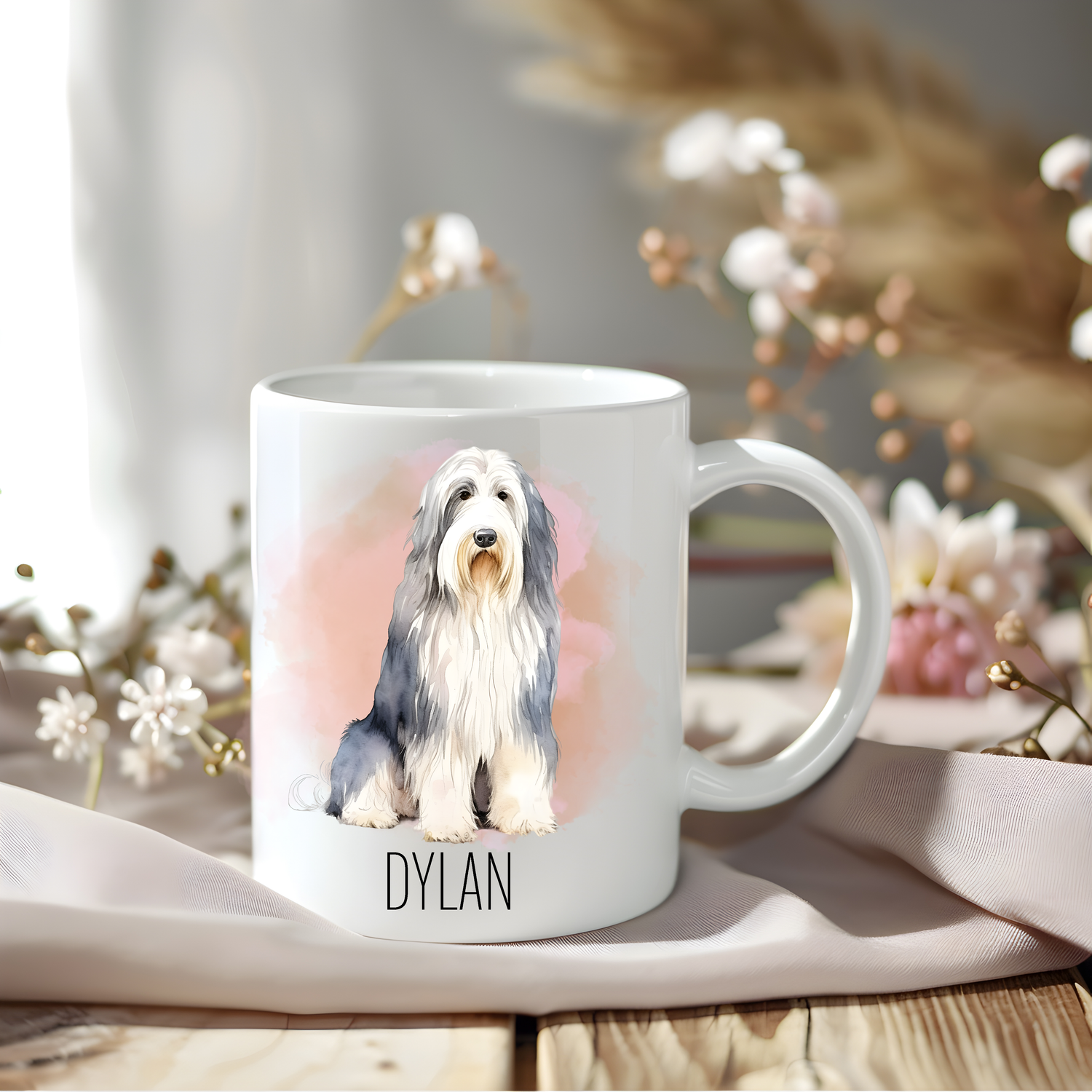 Bearded Collie coffee mug personalised with a pet name.  The mug is sat on a wooden surface with a creamy coloured cloth and various flowers adorn the background. 